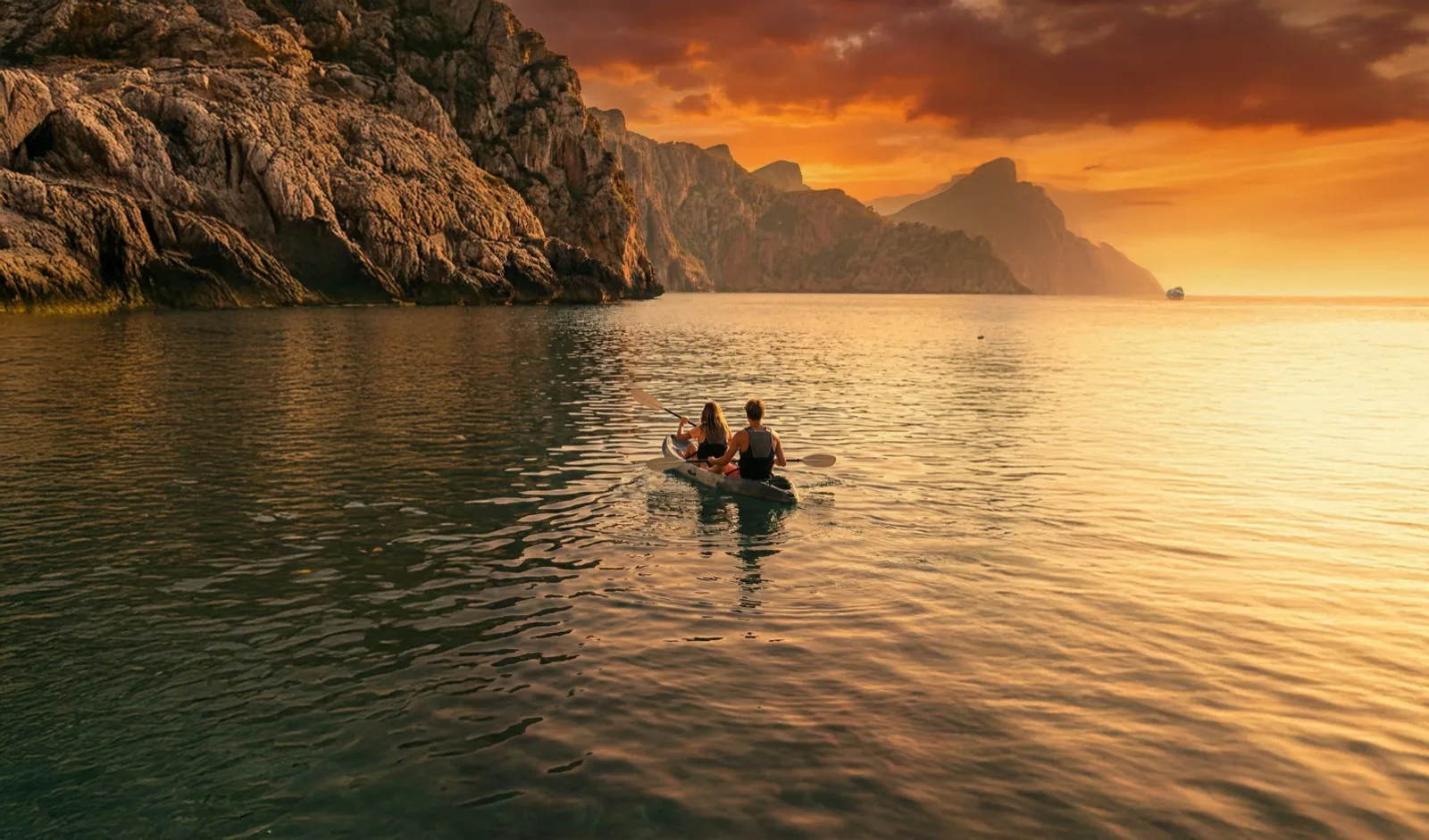 Two people paddle a double kayak at sunset in a Mallorcan cove, with cliffs in the background.
